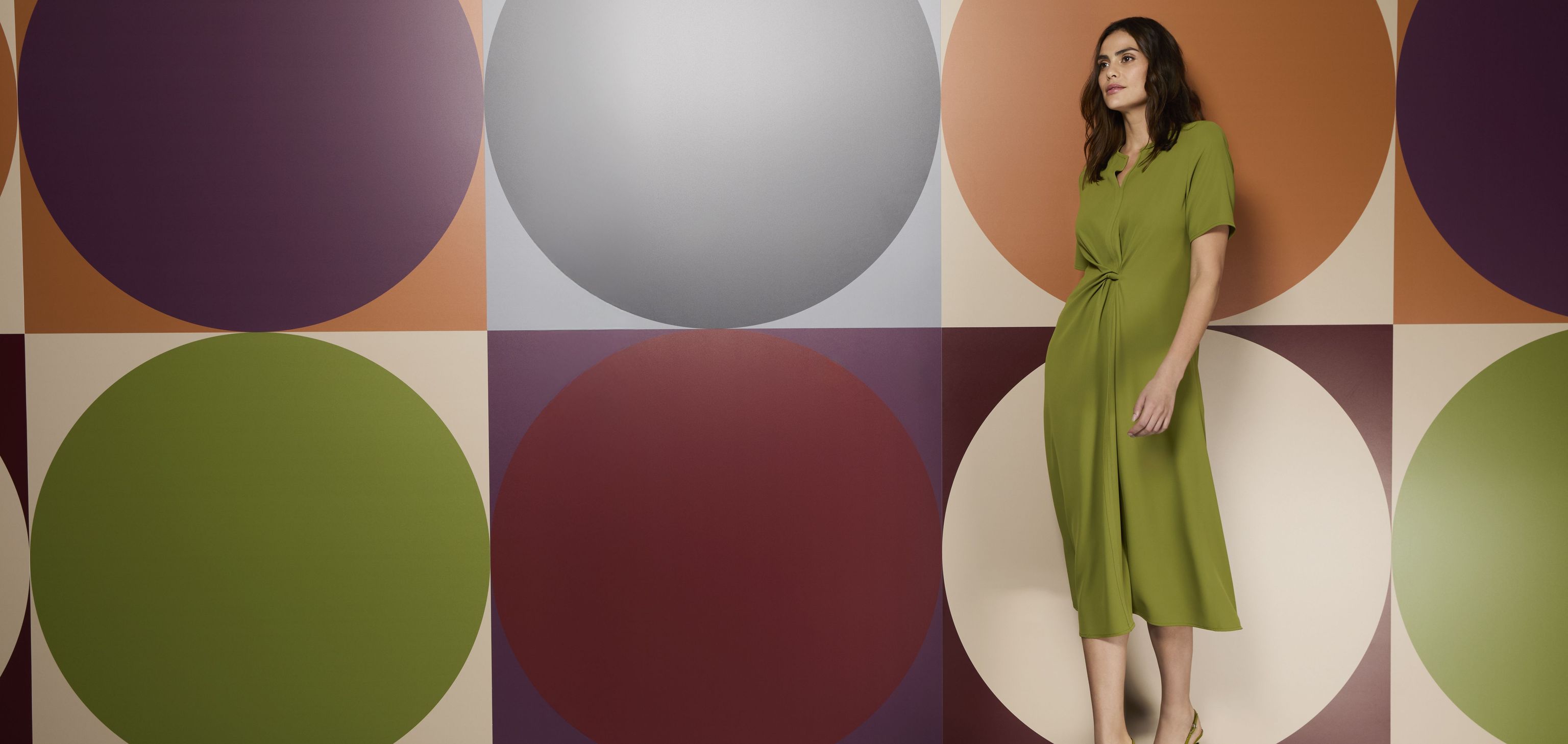 Frau in grünem Kleid vor farbenfrohem Kreis-Muster-Wand, elegant stehend.