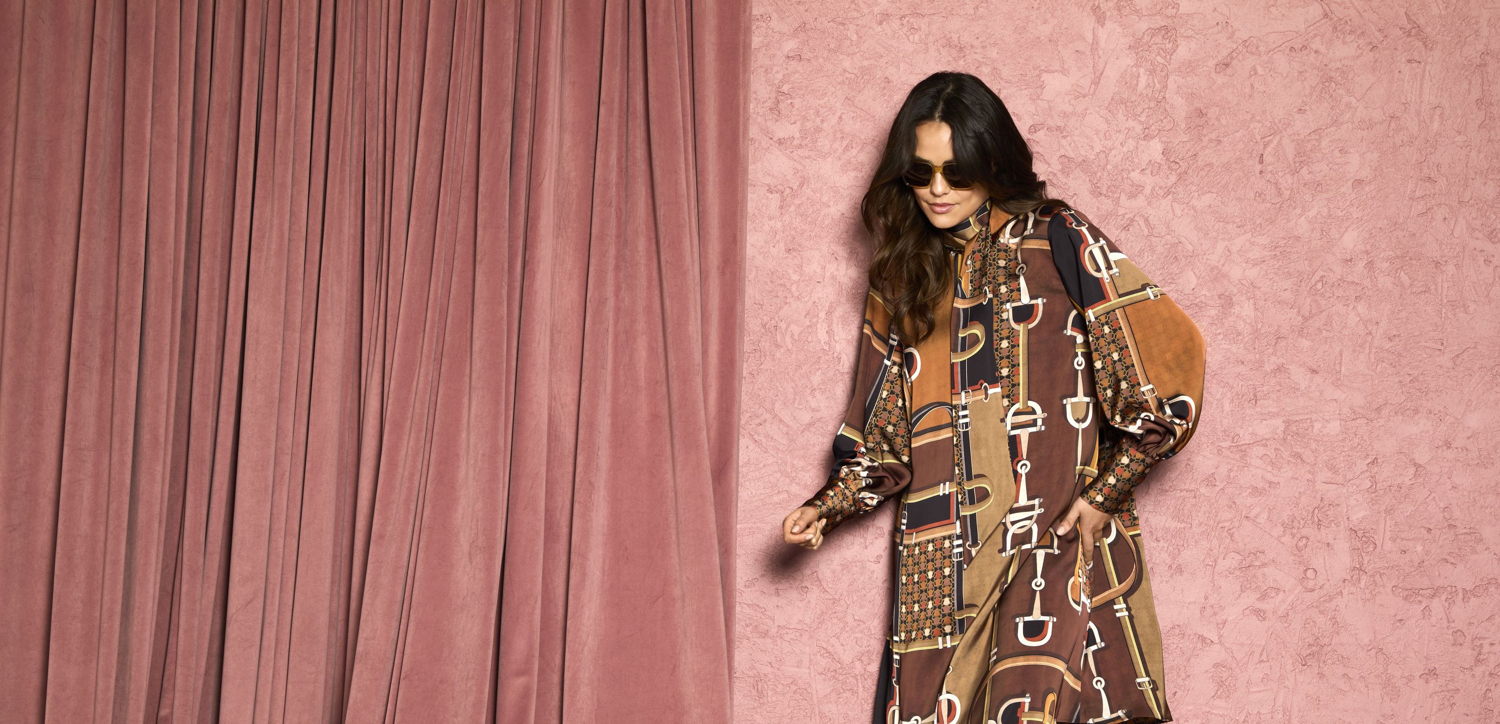 Stylische Person in buntem Patchwork-Kleid mit Riemchen-Accessoires und Stiefeln vor rosafarbenem Vorhang.