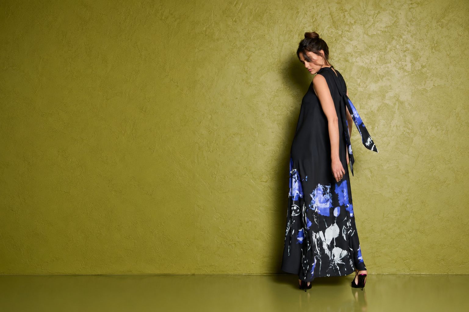 Frau in elegantem, blau-schwarz gemustertem Kleid lehnt lässig an gelber Wand.