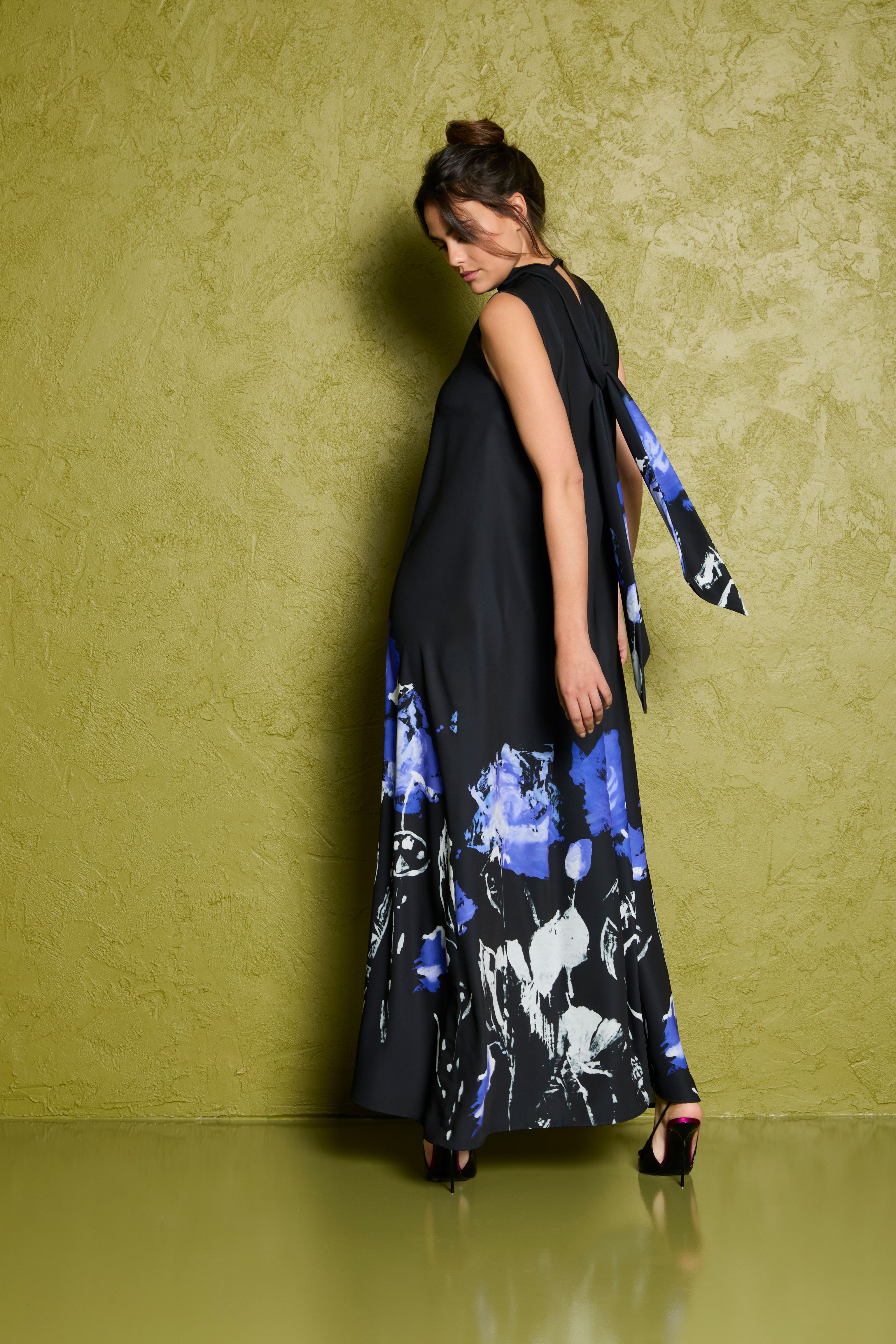 Frau trägt langes, schlichtes Kleid mit blauen floralen Mustern am Saum, lehnt an Wand.
