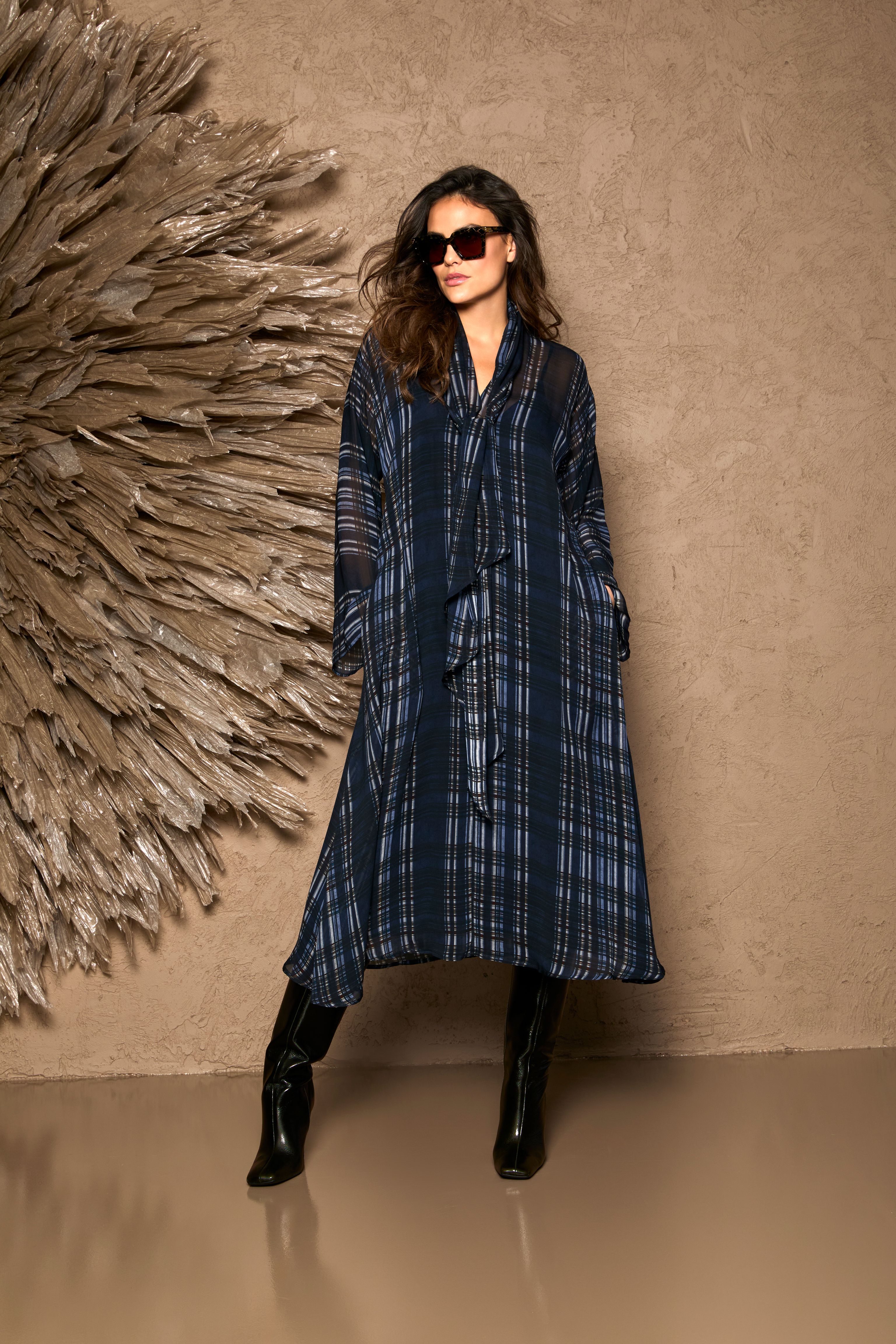 Frau trägt eleganten, langen Plaid-Mantel mit schwarzen Stiefeln und Sonnenbrille.