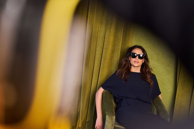 Person mit Sonnenbrille trägt elegantes dunkles Kleid in gelb-grünem Raum.