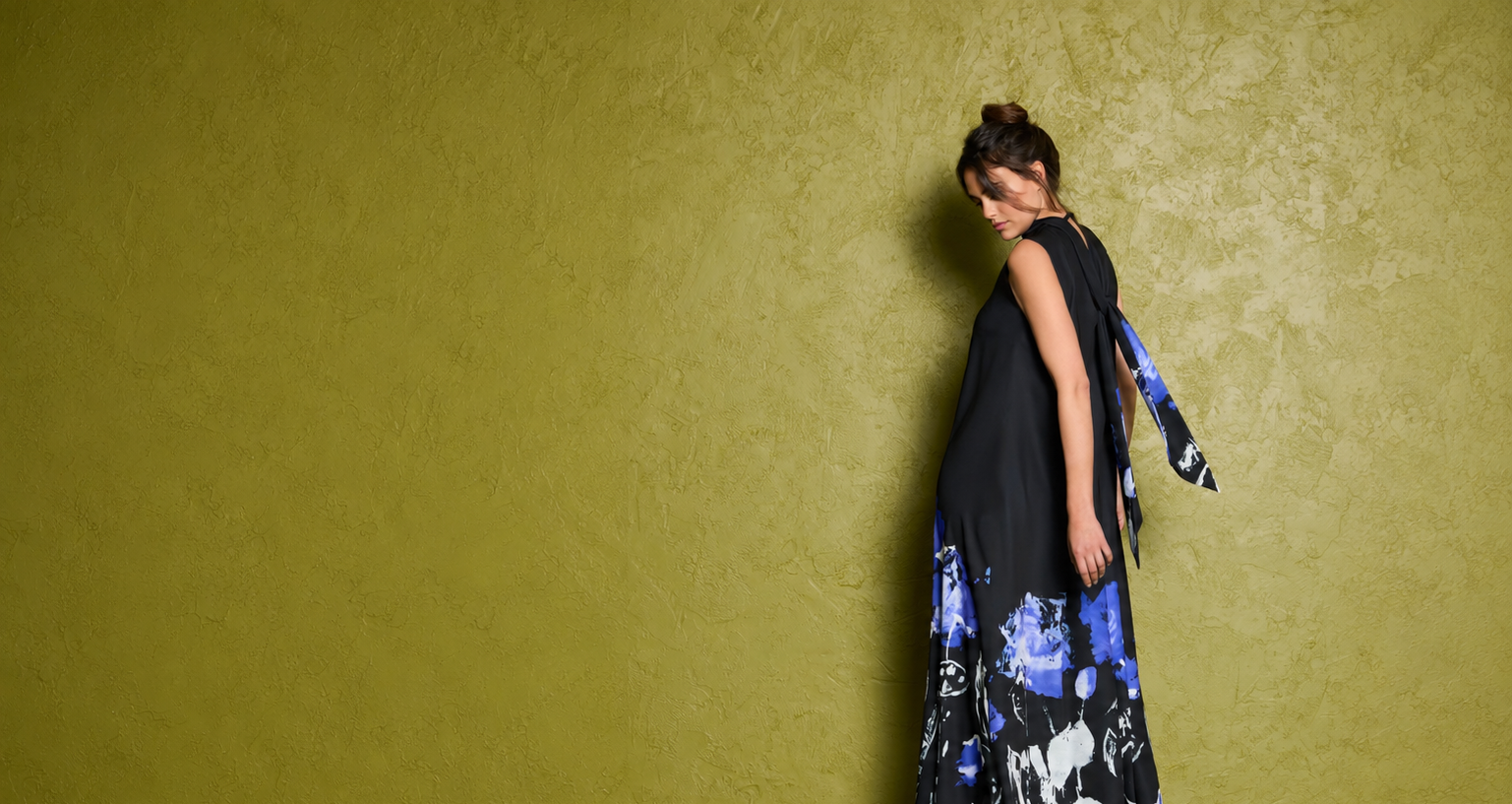 Frau in elegantem, blau-schwarz gemustertem Kleid lehnt an gelber Wand.